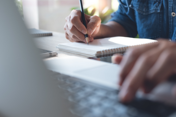 A person at work typing on a laptop and writing on a notepad